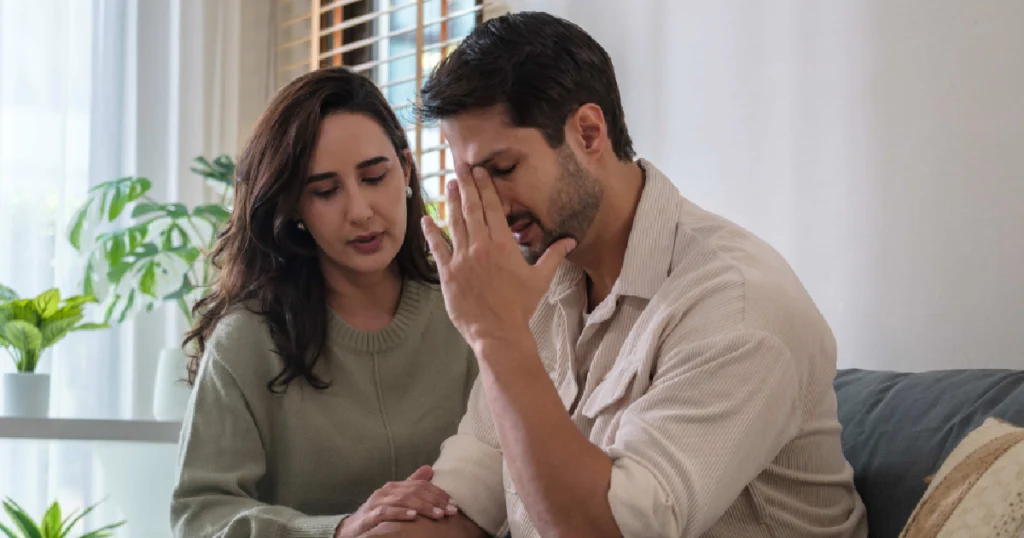A concerned couple sits together on a couch, with the man appearing distressed and holding his head representing emotional struggles related to Low testosterone (Low-T) in Webster, TX.